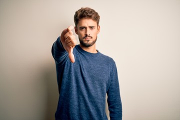 Young handsome man with beard wearing casual sweater standing over white background looking unhappy and angry showing rejection and negative with thumbs down gesture. Bad expression.