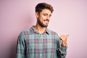 Fototapeta premium Young handsome man with beard wearing casual shirt standing over pink background smiling with happy face looking and pointing to the side with thumb up.