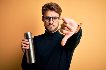 Young man on vacation wearing glasses holding metallic thermo wih water with angry face, negative sign showing dislike with thumbs down, rejection concept