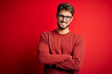 Young handsome man with beard wearing glasses and sweater standing over red background happy face...