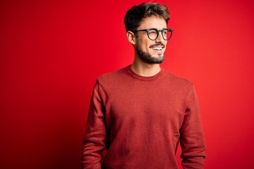 Young handsome man with beard wearing glasses and sweater standing over red background looking away...