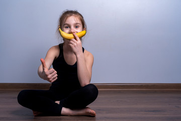 A white-skinned girl in a black sweatshirt holds a banana in her hand and uses it as a smile. The...