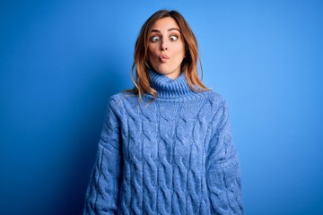 Young beautiful brunette woman wearing casual turtleneck sweater over blue background making fish face with lips, crazy and comical gesture. Funny expression.