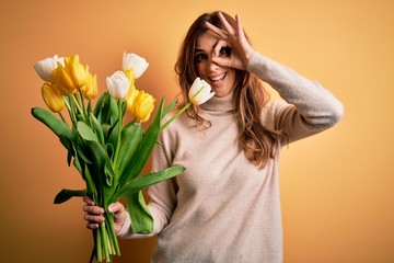 Young beautiful brunette woman holding bouquet of yellow tulips over isolated background doing ok gesture with hand smiling, eye looking through fingers with happy face.