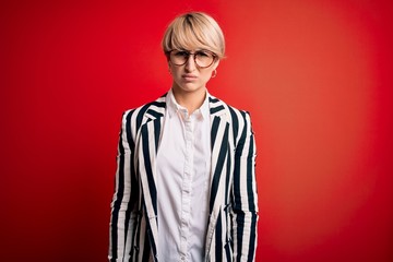 Blonde business woman with short hair wearing glasses and striped jacket over red background skeptic and nervous, frowning upset because of problem. Negative person.