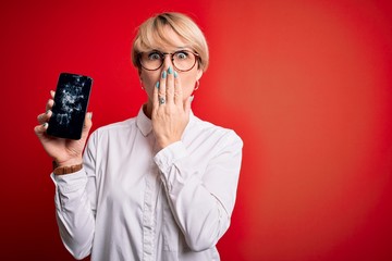 Young blonde woman with short hair holding broken smartphone over red background cover mouth with...