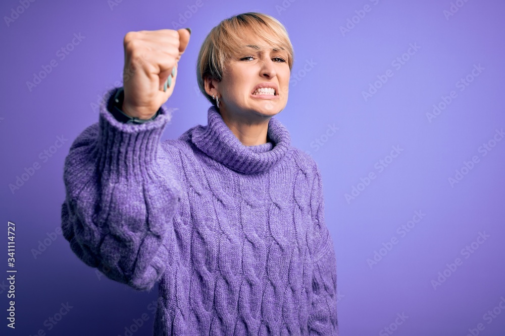 Sticker Young blonde woman with short hair wearing winter turtleneck sweater over purple background angry and mad raising fist frustrated and furious while shouting with anger. Rage and aggressive concept.