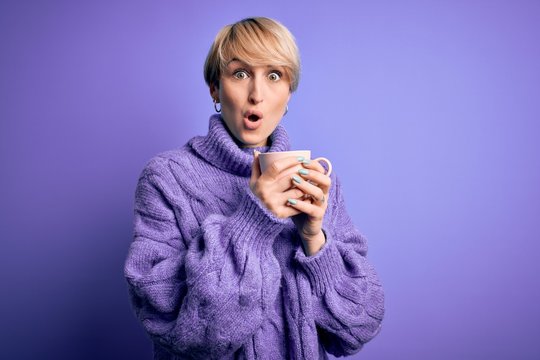 Young blonde woman with short hair wearing winter sweater drinking a cup of hot coffee scared in shock with a surprise face, afraid and excited with fear expression