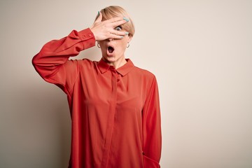 Young beautiful business blonde woman with short hair standing over isolated background peeking in shock covering face and eyes with hand, looking through fingers with embarrassed expression.