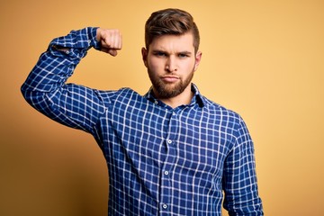 Naklejka premium Young blond businessman with beard and blue eyes wearing shirt over yellow background Strong person showing arm muscle, confident and proud of power