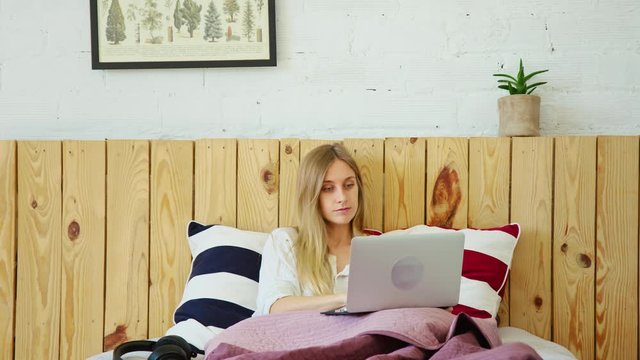 Cute smiling young woman, texts with friends through social media app on her laptop. Woman in bed opens laptop on lazy weekend morning, check notifications