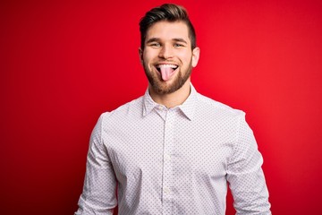 Young blond businessman with beard and blue eyes wearing elegant shirt over red background sticking tongue out happy with funny expression. Emotion concept.