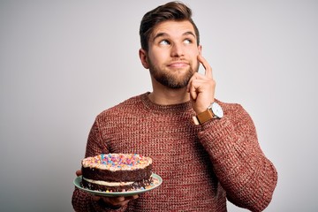 Young blond man with beard and blue eyes holding birthday cake over white background serious face...