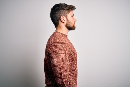 Young blond man with beard and blue eyes wearing casual sweater over white background looking to side, relax profile pose with natural face and confident smile.