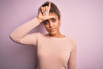 Young beautiful african american girl wearing casual sweater standing over pink background making fun of people with fingers on forehead doing loser gesture mocking and insulting.