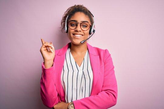 Young african american call center agent girl wearing glasses working using headset with a big smile on face, pointing with hand and finger to the side looking at the camera.