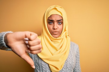 Young beautiful african american girl wearing muslim hijab over isolated yellow background looking unhappy and angry showing rejection and negative with thumbs down gesture. Bad expression.