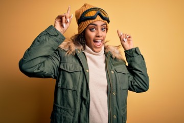 Young african american afro skier girl wearing snow sportswear and ski goggles smiling amazed and surprised and pointing up with fingers and raised arms.