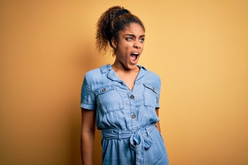 Young beautiful african american girl wearing denim dress standing over yellow background angry and mad screaming frustrated and furious, shouting with anger. Rage and aggressive concept.