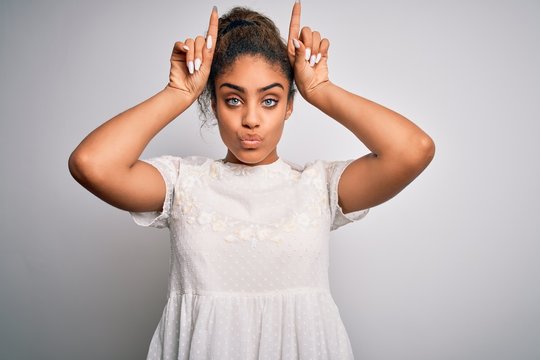 Young beautiful african american girl wearing casual t-shirt standing over white background doing funny gesture with finger over head as bull horns