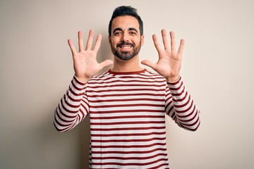 Young handsome man with beard wearing casual striped t-shirt standing over white background showing and pointing up with fingers number ten while smiling confident and happy.