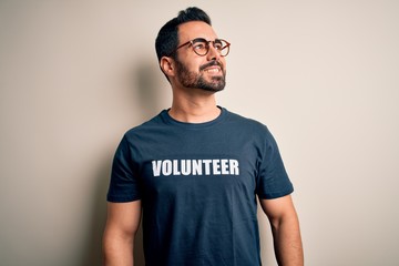 Handsome man with beard wearing t-shirt with volunteer message over white background looking away to side with smile on face, natural expression. Laughing confident.