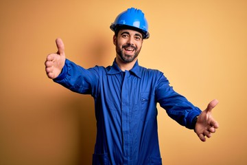 Mechanic man with beard wearing blue uniform and safety helmet over yellow background looking at the camera smiling with open arms for hug. Cheerful expression embracing happiness.