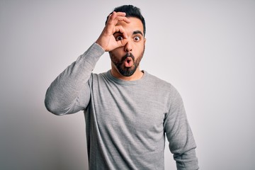 Young handsome man with beard wearing casual sweater standing over white background doing ok gesture shocked with surprised face, eye looking through fingers. Unbelieving expression.