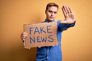 Young handsome redhead man holding banner with fake news message over yellow background with open...