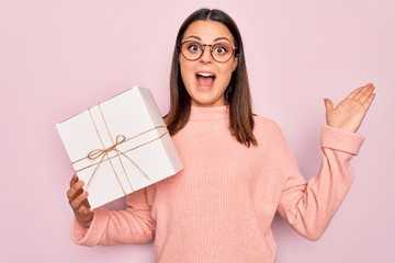 Young beautiful brunette woman holding birthday gift over isolated pink background celebrating victory with happy smile and winner expression with raised hands