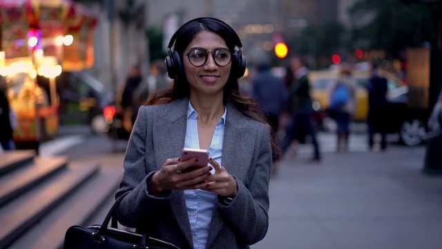 South Asian woman in optical spectacles and bluetooth electronic earphones walking around city streets and smiling while listening motivation audio book from cellphone app, slow motion effect
