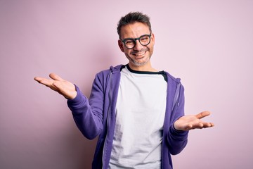 Young handsome man wearing purple sweatshirt and glasses standing over pink background smiling cheerful with open arms as friendly welcome, positive and confident greetings