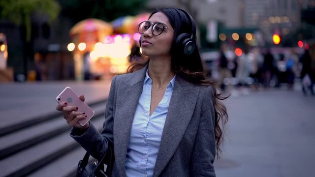 South Asian business woman formally dressed walking at urban street with smartphone device in hand and smiling, slow motion effect successful female manager in eyewear enjoying music time
