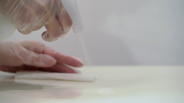 Woman in protective gloves cleaning white table with cloth and spray.
