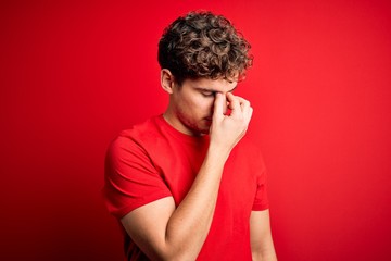 Young blond handsome man with curly hair wearing casual t-shirt over red background tired rubbing nose and eyes feeling fatigue and headache. Stress and frustration concept.
