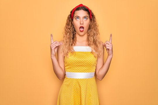 Beautiful blonde pin-up woman with blue eyes wearing diadem standing over yellow background amazed and surprised looking up and pointing with fingers and raised arms.
