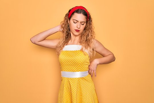 Beautiful blonde pin-up woman with blue eyes wearing diadem standing over yellow background Suffering of neck ache injury, touching neck with hand, muscular pain