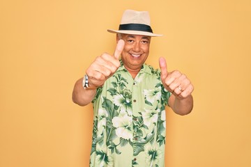 Middle age senior grey-haired man wearing summer hat and floral shirt on beach vacation approving doing positive gesture with hand, thumbs up smiling and happy for success. Winner gesture.