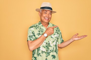 Middle age senior grey-haired man wearing summer hat and floral shirt on beach vacation amazed and smiling to the camera while presenting with hand and pointing with finger.