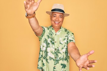 Middle age senior grey-haired man wearing summer hat and floral shirt on beach vacation looking at the camera smiling with open arms for hug. Cheerful expression embracing happiness.