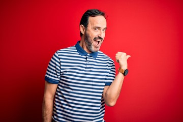 Fototapeta premium Middle age hoary man wearing casual striped polo standing over isolated red background smiling with happy face looking and pointing to the side with thumb up.