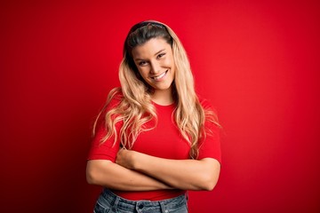 Young beautiful blonde woman wearing casual t-shirt standing over isolated red background happy face smiling with crossed arms looking at the camera. Positive person.