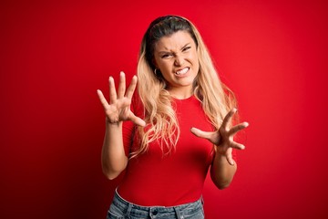 Young beautiful blonde woman wearing casual t-shirt standing over isolated red background smiling funny doing claw gesture as cat, aggressive and sexy expression