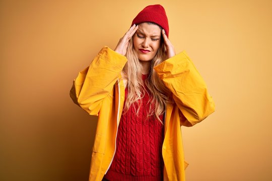 Young beautiful blonde woman wearing raincoat and wool cap over isolated yellow background suffering from headache desperate and stressed because pain and migraine. Hands on head.