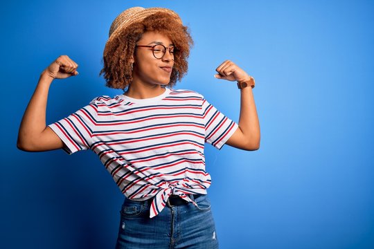 African American woman with curly hair on vacation wearing summer hat and striped t-shirt showing arms muscles smiling proud. Fitness concept.