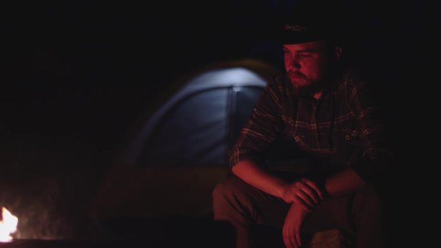 Man in hat sits by a campfire at night in slow motion