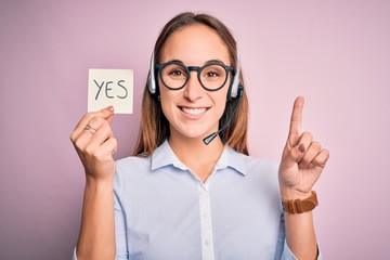 Beautiful call center agent woman working using headset holding reminder with yes message surprised...