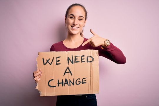Young beautiful activist woman asking for change protesting holding banner with message with surprise face pointing finger to himself