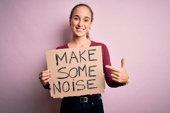 Beautiful activist woman holding banner with make some noise message over pink background very happy pointing with hand and finger
