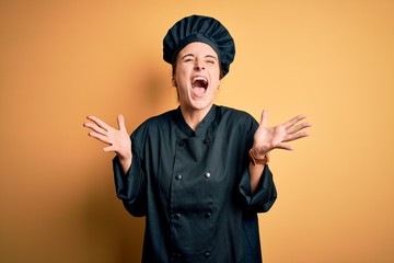 Young beautiful chef woman wearing cooker uniform and hat standing over yellow background celebrating mad and crazy for success with arms raised and closed eyes screaming excited. Winner concept